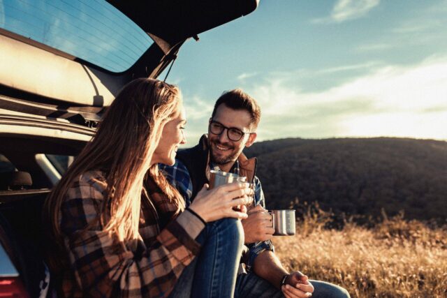 A couple sitting on their car boot in nature, drinking a hot drink