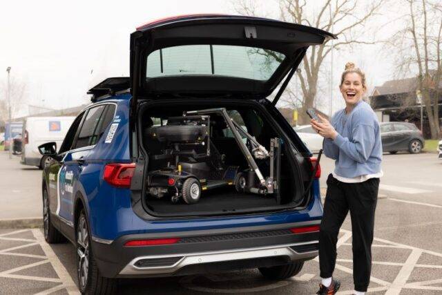 Paralympian Robyn Love stood at the open boot of a car holding the handset for the Boot Hoist. There is a mobility scooter loaded into the boot of the car.