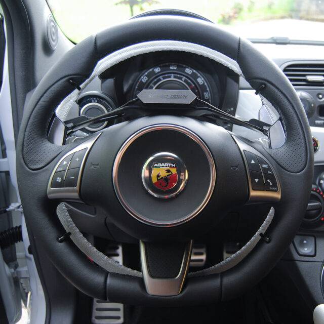 A driver's seat view of the steering wheel. An Underer Ring accelerator control for a car, a thin black ring connected by a control box, is attached behind the steering wheel.
