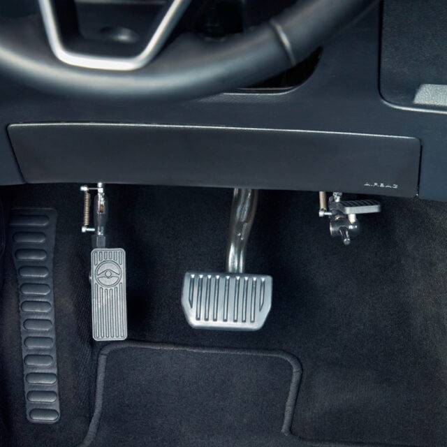 A driver's seat view of a car interior. In the footwell the accelerator is folded up and out of the way, a second accelerator pedal has been folded down to the left of the accelerator pedal.