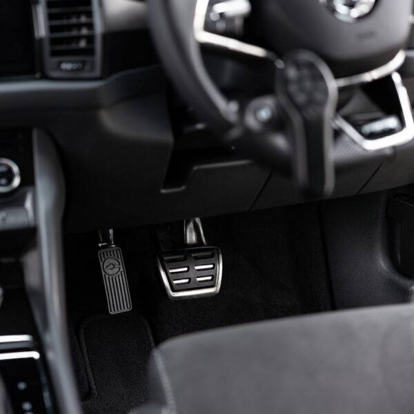 A driver's seat view of a car interior. There is a steering aid on the steering whee. In the footwell the accelerator is folded up and out of the way, a second accelerator pedal has been folded down to the left of the accelerator pedal.