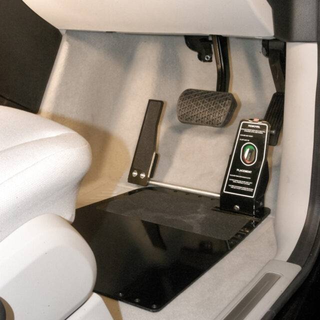 A driver's seat view of a car interior. In the footwell, there is a metal plate on the floor supporting a guard covering the accelerator pedal. Attached to the guard is a second accelerator pedal positioned on the left-hand side of the brake pedal.