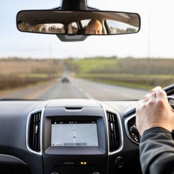 The interior of a car showing a panoramic rear view mirror attached over the top of the car's original mirror. It is wider than a traditional rear view mirror. The reflection in the mirror shows more of the car than the original mirror could.