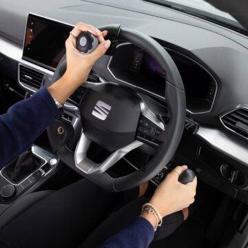 A driver's seat view of the wheel in an adapted car with mechanical push/pull hand controls next to the steering wheel and a steering ball