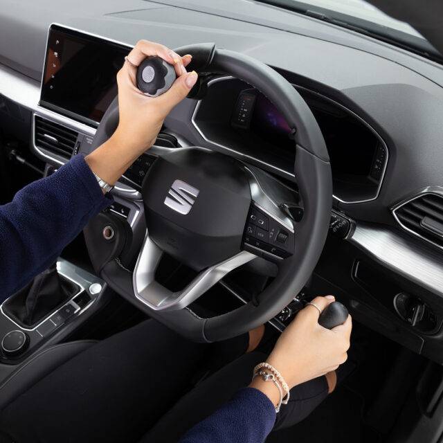 A driver's seat view of the wheel in an adapted car with mechanical push/pull hand controls next to the steering wheel and a steering ball