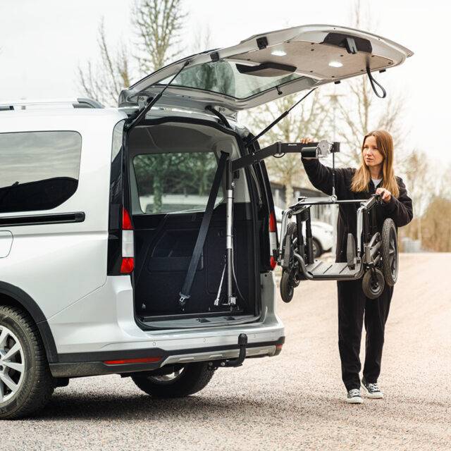 A young woman stood beside the open boot of her car with the Carony wheelchair base from a swivel seat. It is being lifted out of the car boot by the BraunAbility Carolift 40 hinged boot hoist.
