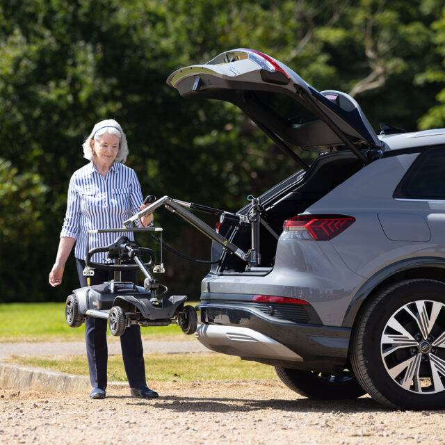 An older woman stood beside the open boot of her car with a mobility scooter. It is being lifted out of the car boot by the Autochair Smart Lifter LC compact Boot Hoist.