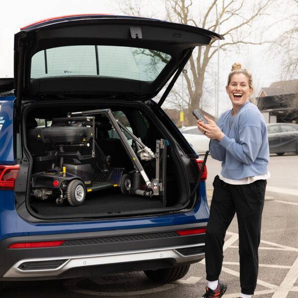 Paralympian Robyn Love stood at the open boot of a car holding the handset for the Boot Hoist. There is a mobility scooter loaded into the boot of the car.