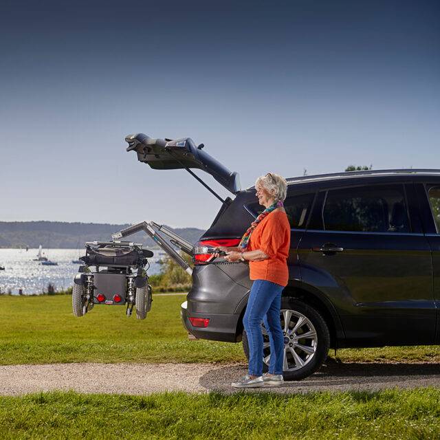 An older woman stood beside the open boot of her car with a large power chair. It is being lifted out of the car boot by the Autochair Smart Lifter LP Boot Hoist.