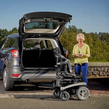 An older woman stood beside the open boot of her car with a large power chair. It has been lifted out of the car boot by the Autochair Smart Lifter LP Boot Hoist.
