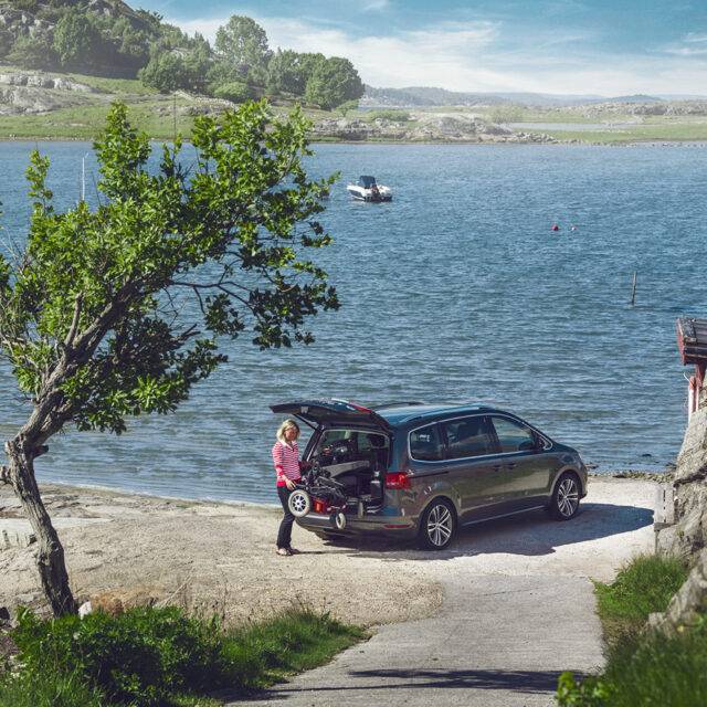 A woman stood beside the open boot of her car with a power chair. It is being lifted out of the car boot by the BraunAbility Carolift 6900 Boot Hoist.