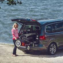 A woman stood beside the open boot of her car with a power chair. It is being lifted out of the car boot by the BraunAbility Carolift 6900 Boot Hoist.