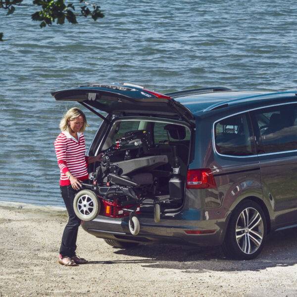 A woman stood beside the open boot of her car with a power chair. It is being lifted out of the car boot by the BraunAbility Carolift 6900 Boot Hoist.
