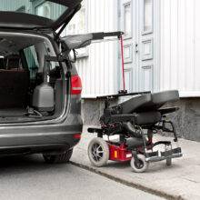 The open boot of a car with a large power chair beside it on the pavement. It is being lifted out of the car boot by the BraunAbility Carolift 6900 Boot Hoist thanks to its hinged telescopic 6-way arm.