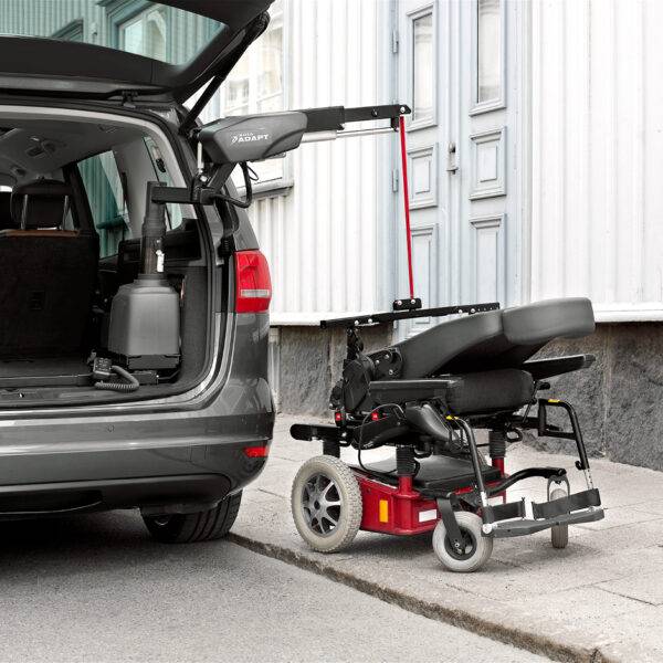 The open boot of a car with a large power chair beside it on the pavement. It is being lifted out of the car boot by the BraunAbility Carolift 6900 Boot Hoist thanks to its hinged telescopic 6-way arm.