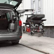 The open boot of a car with a large power chair beside it above the pavement. It is being lifted out of the car boot by the BraunAbility Carolift 6900 Boot Hoist thanks to its hinged telescopic 6-way arm.