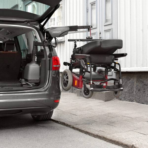 The open boot of a car with a large power chair beside it above the pavement. It is being lifted out of the car boot by the BraunAbility Carolift 6900 Boot Hoist thanks to its hinged telescopic 6-way arm.