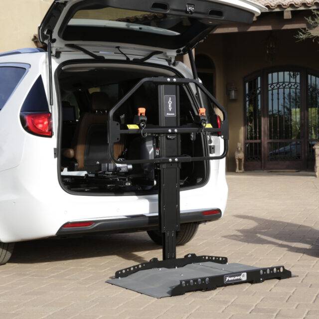 The open boot of a car with the Joey Platform Lift from Bruno. A black and grey metal platform for raising and loading a mobility device into the boot of a vehicle. It had a robust frame a wide adjustable platform.