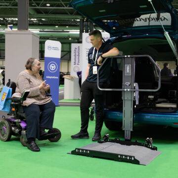 A woman in a powerchair speaking to an assessor from Mobility in Motion next to the boot of a car with the Joey Platform Lift from Bruno.