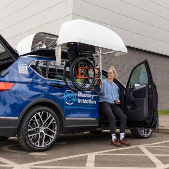 Paralympian Robyn Love sat on the Autochair Transfer Plate beside the driver's seat of the car, bringing her outside the door frame of the car to more easily reach the wheelchair being lowered down to her by the BraunAbility Chair Topper wheelchair roof box.