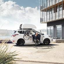 A man sat on the Turny Evo swivel seat has turned the seat out of the car and lowered it down to bring him closer to the wheelchair for an easier transfer at the ideal height. The wheelchair is being lowered down to him by the BraunAbility Chair Topper wheelchair roof box.