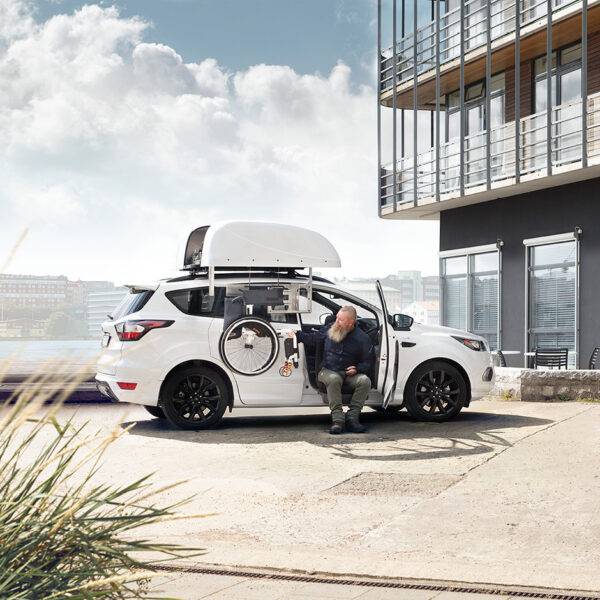 A man sat on the Turny Evo swivel seat has turned the seat out of the car and lowered it down to bring him closer to the wheelchair for an easier transfer at the ideal height. The wheelchair is being lowered down to him by the BraunAbility Chair Topper wheelchair roof box.