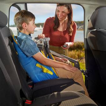 A child is being lifted comfortably into the rear passenger seat of the car in a sling. His mother is using the Autochair Milford Person Lift, a smart grey person hoist with a blue sling, to lift the boy up and into the car.