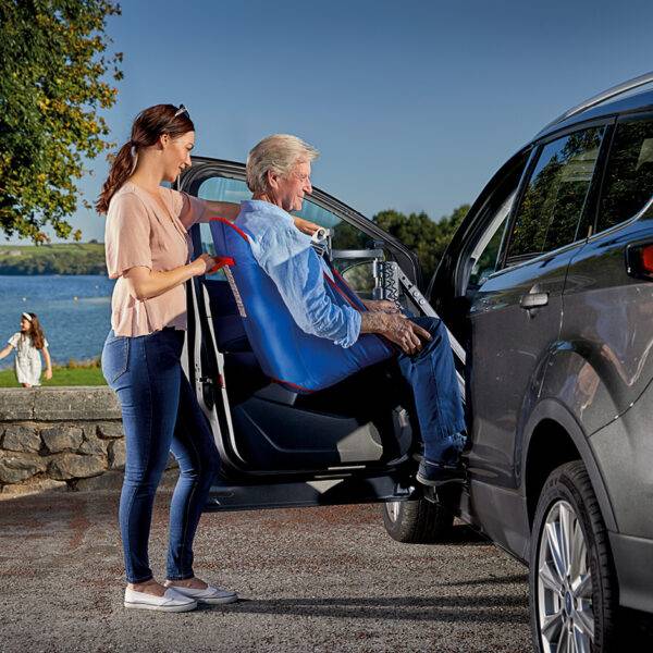 An older man being lifted comfortably into the passenger seat of the car in a sling. A young woman is using the Autochair Milford Person Lift, a smart grey person hoist with a blue sling, to lift the man up and into the car.