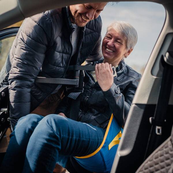 An older woman being lifted comfortably into the driver's seat of the car in a sling. A young man is using the Autochair Milford Person Lift, a smart grey person hoist with a blue sling, to lift the man up and into the car.