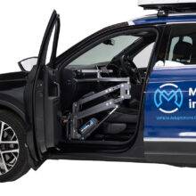The open passenger door of a car reveals the lifting arm of the Autochair Milford Person Lift, a smart grey person hoist with a blue sling, to lift occupants safely into the original seat of the car.