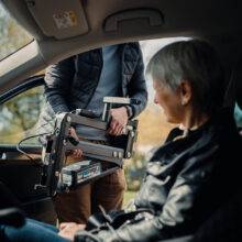 An older woman sat comfortably in the driver's seat of the car in the sling of the Milford Person Lift. A young man is removing the lifting arm of the person hoist so the door can be closed for their journey.