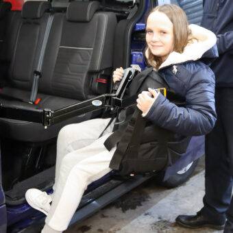 A child is being lifted comfortably into the rear passenger seat of the car in a sling. A young man is using the BAS Person Lift, a versatile second-row person hoist with a black hinged lifting arm and a black sling, to lift the girl up and into the car.