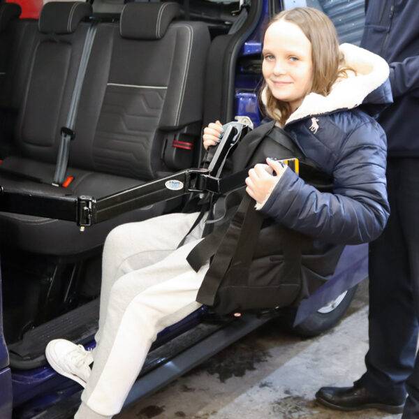 A child is being lifted comfortably into the rear passenger seat of the car in a sling. A young man is using the BAS Person Lift, a versatile second-row person hoist with a black hinged lifting arm and a black sling, to lift the girl up and into the car.