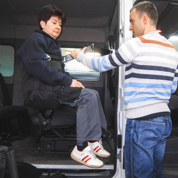 A woman is being lifted comfortably into the rear passenger seat of the car in a sling. A young man is using the BAS Person Lift, a versatile second-row person hoist with a black hinged lifting arm and a black sling, to lift the woman up and into the car.