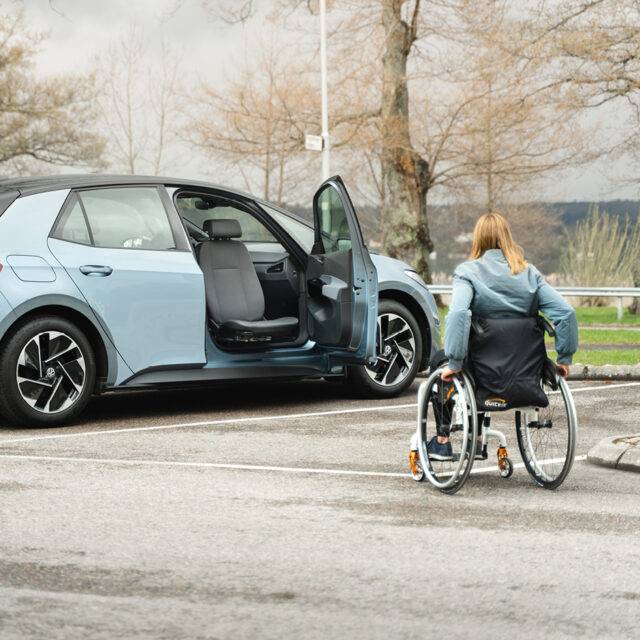 A young woman in a wheelchair approaching her car. The driver's door is open and the swivel seat is turned out of the car to make her transfer from her wheelchair into the car easier.