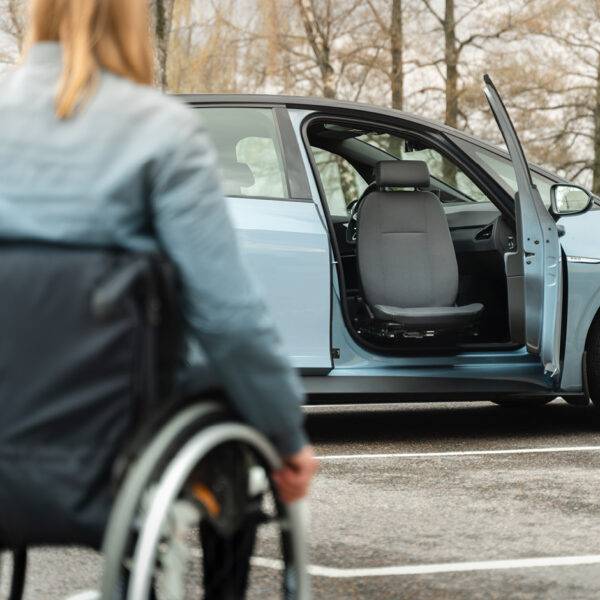 A young woman in a wheelchair approaching her car. The driver's door is open and the swivel seat is turned out of the car to make her transfer from her wheelchair into the car easier.