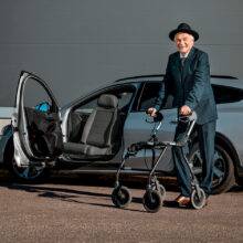 An older man with a mobility frame stood beside his car. A Turny Low swivel seat has turned the seat out of the car to bring the chair closer to the man to make it easier for him to get into the car.