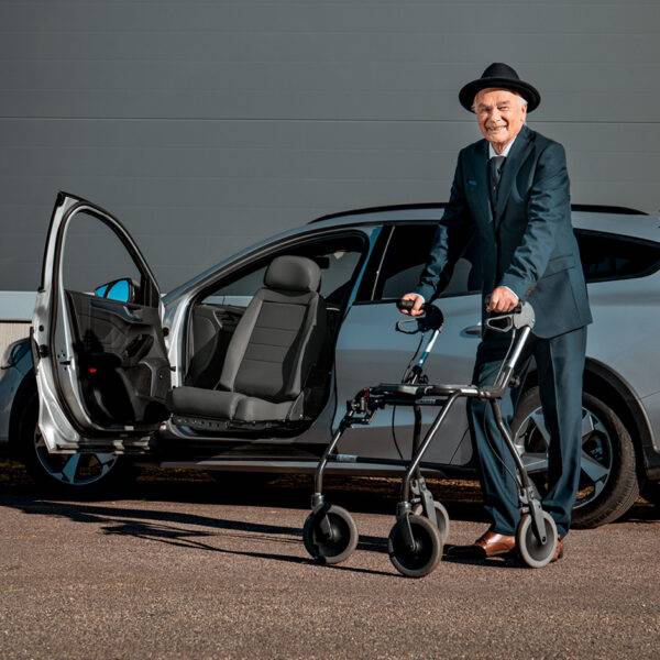 An older man with a mobility frame stood beside his car. A Turny Low swivel seat has turned the seat out of the car to bring the chair closer to the man to make it easier for him to get into the car.