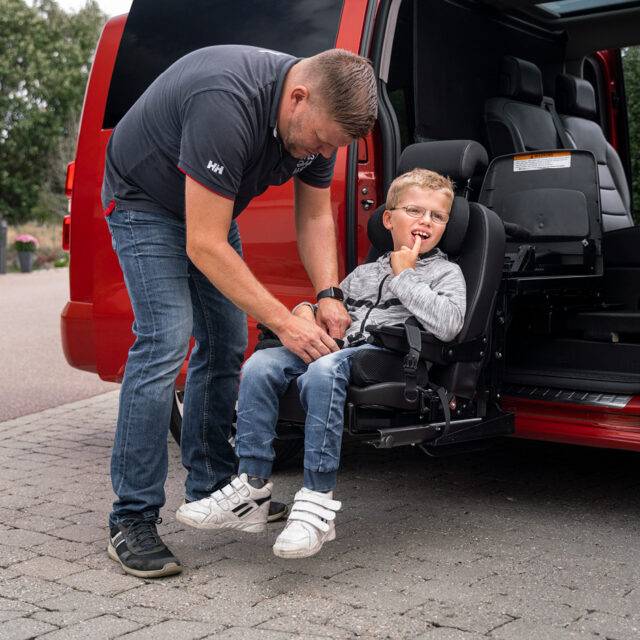 A father helping his son into a car with a swivel seat. The mini-van style vehicle has its passenger sliding door open. The seat has turned out of the car and lowered down to a height to allow the child to easily access it. Additional padding on the seat makes it the right size for a small child.