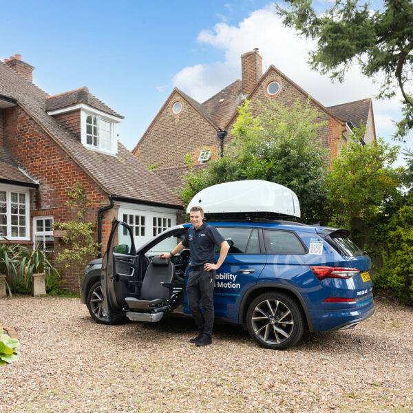 A Mobility in Motion assessor stands next to their demonstration vehicle. The passenger's door is open and a swivel seat has turned out and lowered down to allow for easier access to the chair
