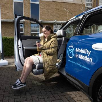 A woman sits in a swivel seat that has turned out of the passenger's seat and lowered outside of the car. The passenger's seat has turned out of the car and lowered down to a height to allow for easier access to the vehicle