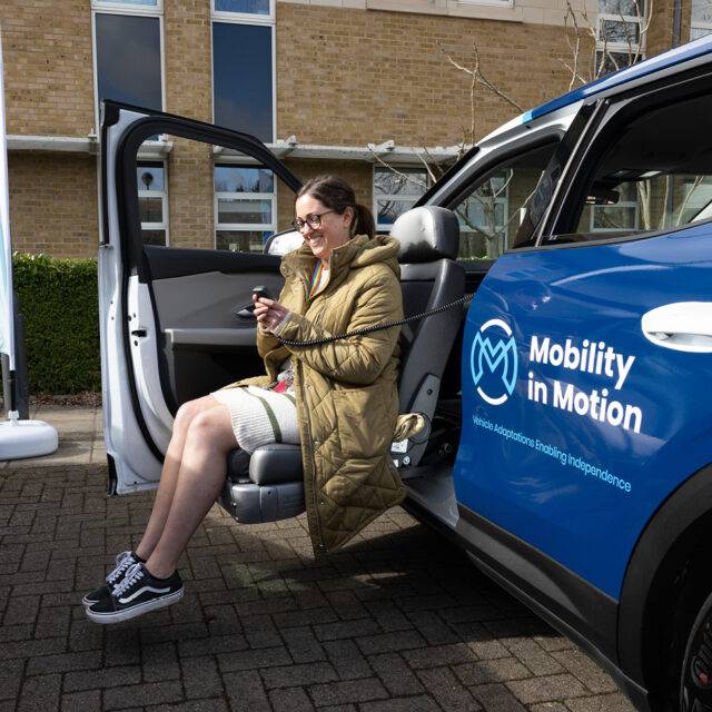 A woman sits in a swivel seat that has turned out of the passenger's seat and lowered outside of the car. The passenger's seat has turned out of the car and lowered down to a height to allow for easier access to the vehicle