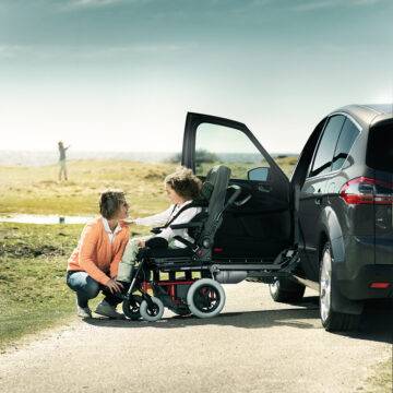 A mother helping his daughter out of a car with a wheelchair swivel seat. The passenger seat has turned out of the car and lowered down to a height to allow the seat to slide off the swivel mechanism and onto a waiting set of wheels that will turn the car seat into a wheelchair. Additional padding on the seat makes it the right size for the small child.
