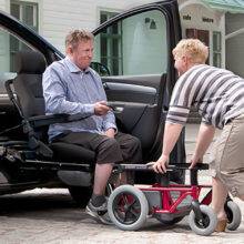 A woman helping a man out of a car with a wheelchair swivel seat. The driver's seat has turned out of the car and lowered down to a height to allow the seat to slide off the swivel mechanism and onto a waiting set of wheels that will turn the car chair into a wheelchair.