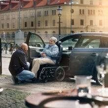 A son helping his elderly mother out of a car with a wheelchair swivel seat. The passenger seat has turned out of the car to allow the seat to slide off the swivel mechanism and onto a waiting set of wheels that will turn the car chair into a wheelchair.
