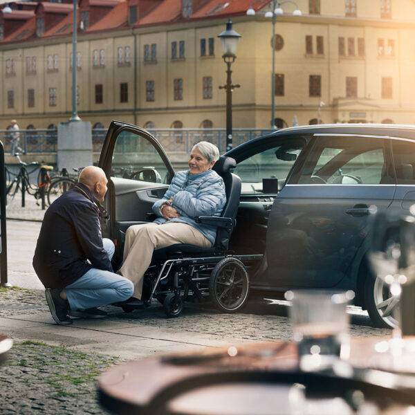 A son helping his elderly mother out of a car with a wheelchair swivel seat. The passenger seat has turned out of the car to allow the seat to slide off the swivel mechanism and onto a waiting set of wheels that will turn the car chair into a wheelchair.