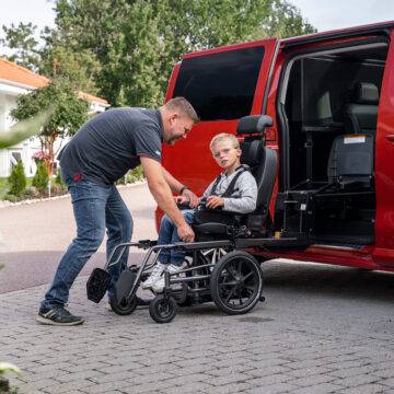 A father helping his son out of a car with a wheelchair swivel seat. The mini-van style vehicle has its passenger sliding door open. The seat has turned out of the car and lowered down to a height to allow the seat to slide off the swivel mechanism and onto a waiting set of wheels that will turn the car chair into a wheelchair. Additional padding on the seat makes it the right size for a small child.