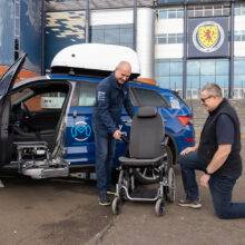 A Mobility in Motion assessor shows a Carony System wheelchair swivel seat to an Occupational Therapist. The car's passenger door is open and the swivel mechanism has turned out and lowered down. Separate from the vehicle, the seat has been transferred onto a wheelchair base.