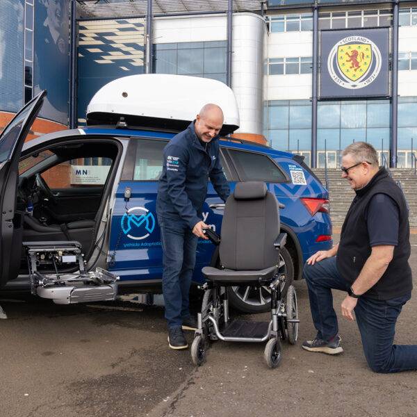 A Mobility in Motion assessor shows a Carony System wheelchair swivel seat to an Occupational Therapist. The car's passenger door is open and the swivel mechanism has turned out and lowered down. Separate from the vehicle, the seat has been transferred onto a wheelchair base.