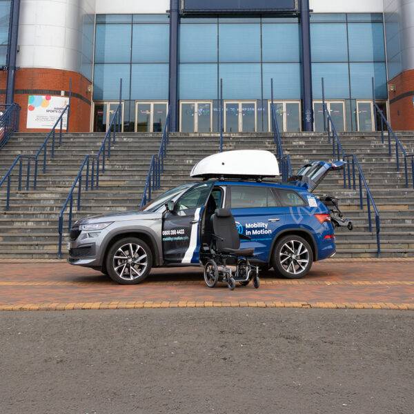 A Mobility in Motion demonstration car with a wheelchair roof box, a boot hoist and a Carony System wheelchair swivel seat. The car's passenger door is open and the swivel mechanism has turned out and lowered down. Separate from the vehicle, the seat has been transferred onto a wheelchair base.